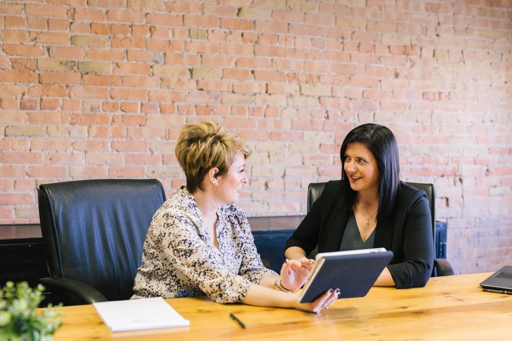 Two woman talking in an office space. One woman is holding a tablet.