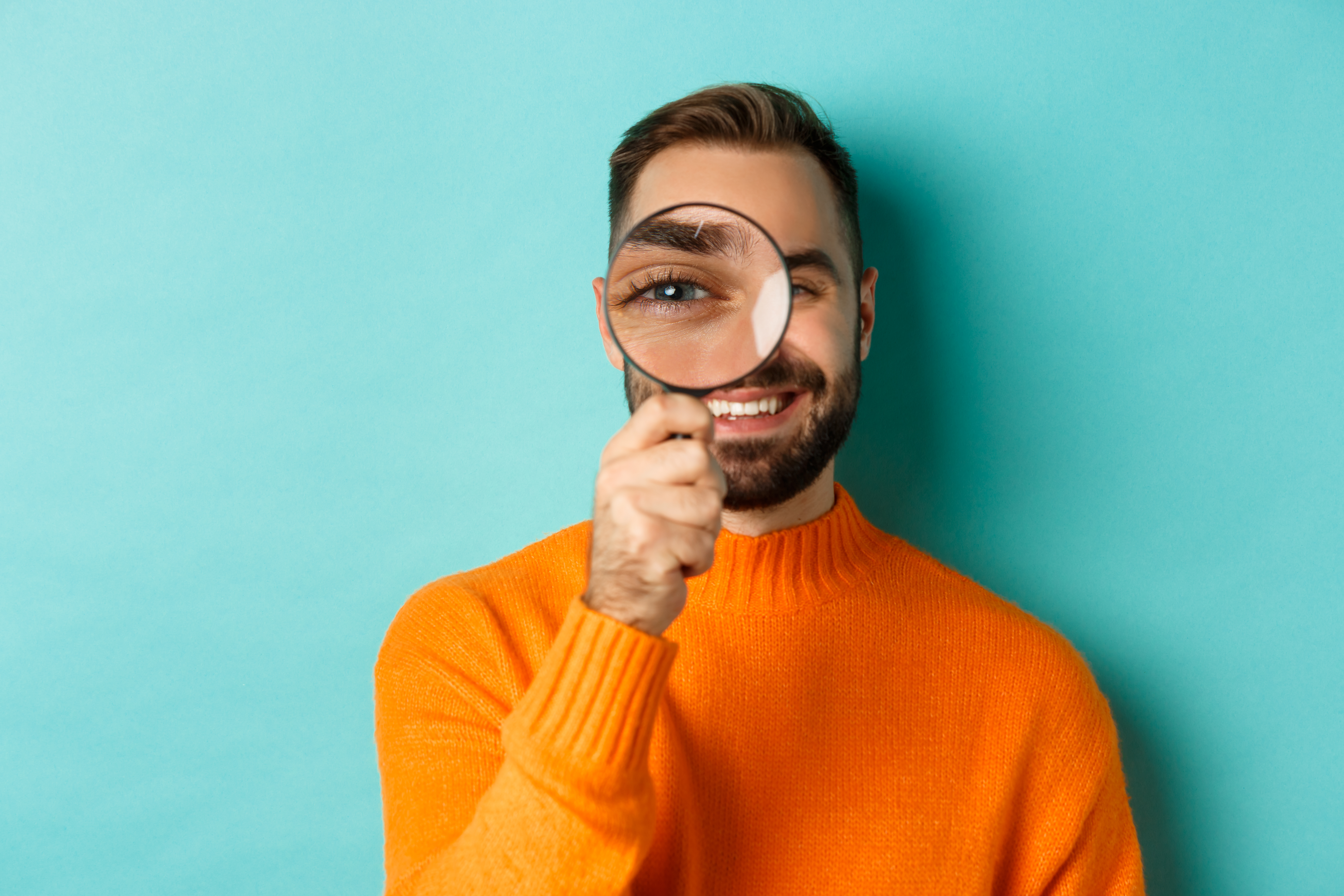 Funny man looking through magnifying glass, searching or investigating something, standing in orange sweater against turquoise background.
