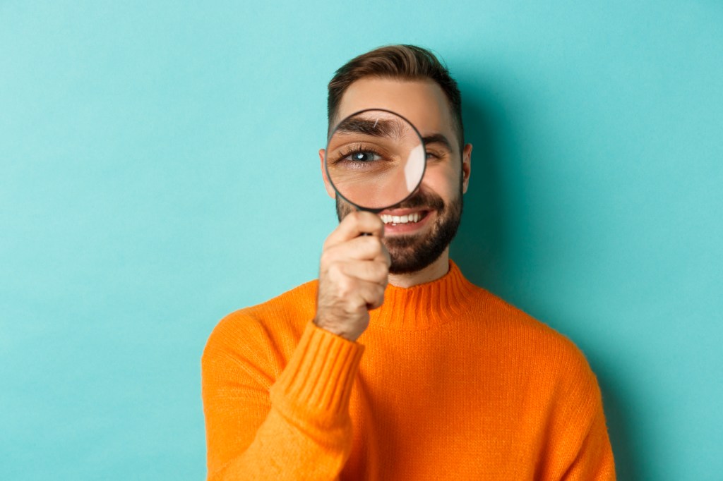 Funny man looking through magnifying glass, searching or investigating something, standing in orange sweater against turquoise background.