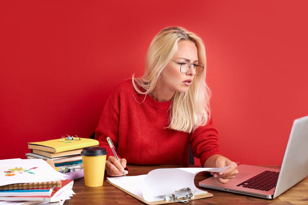 Woman concentrating on work on laptop, she is looking at screen, thinking and write information on paper.