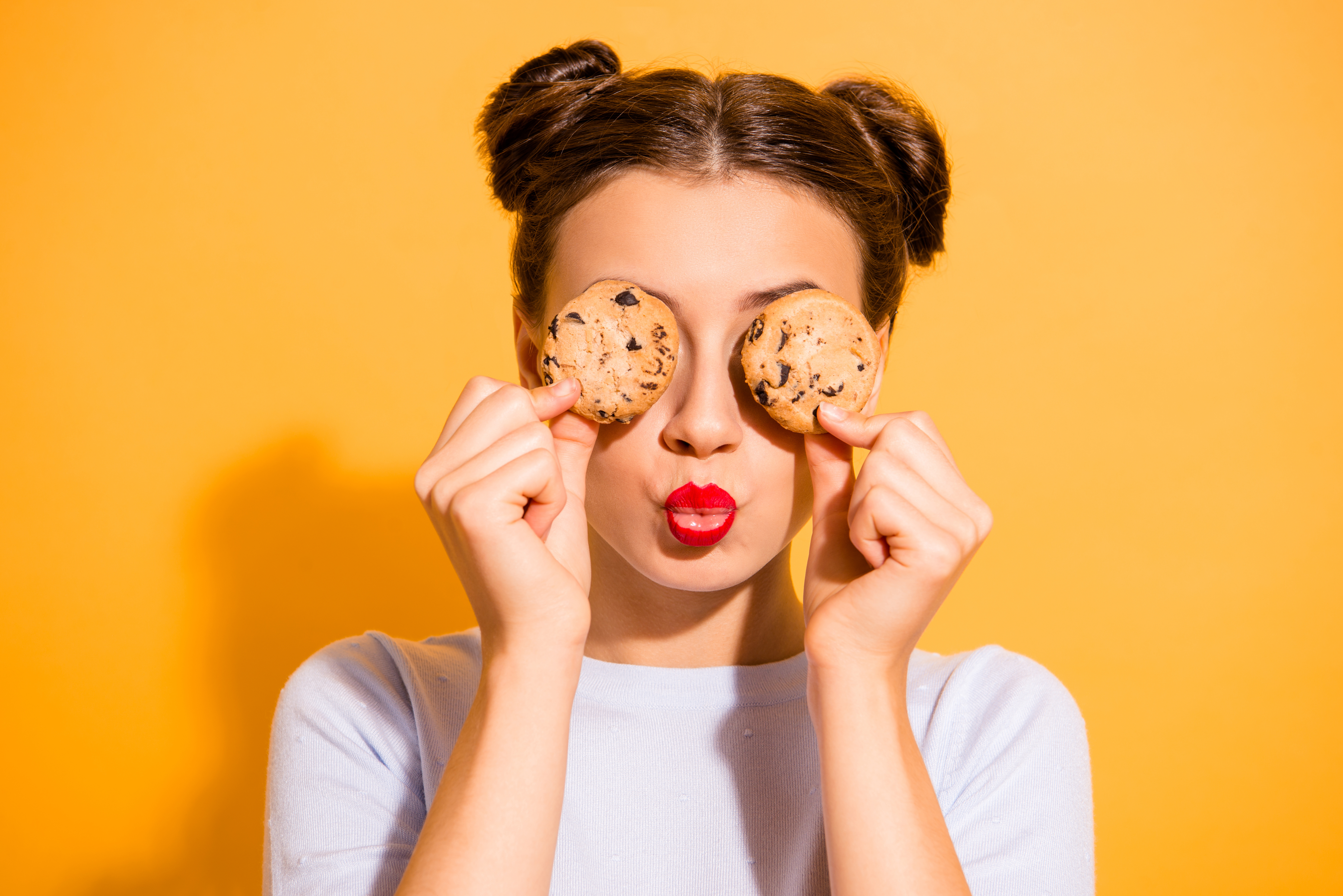 Woman hiding her eyes with cookies dressed in white top isolated over vibrant gold background.