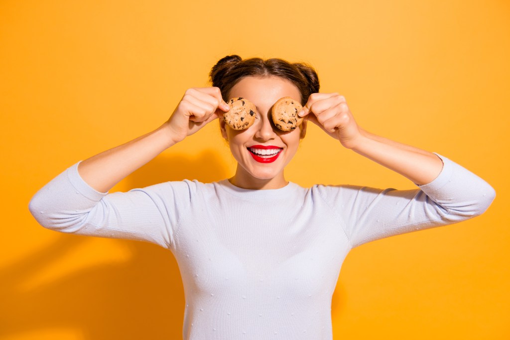 Woman hiding her eyes with cookies dressed in white top isolated over vibrant gold background.