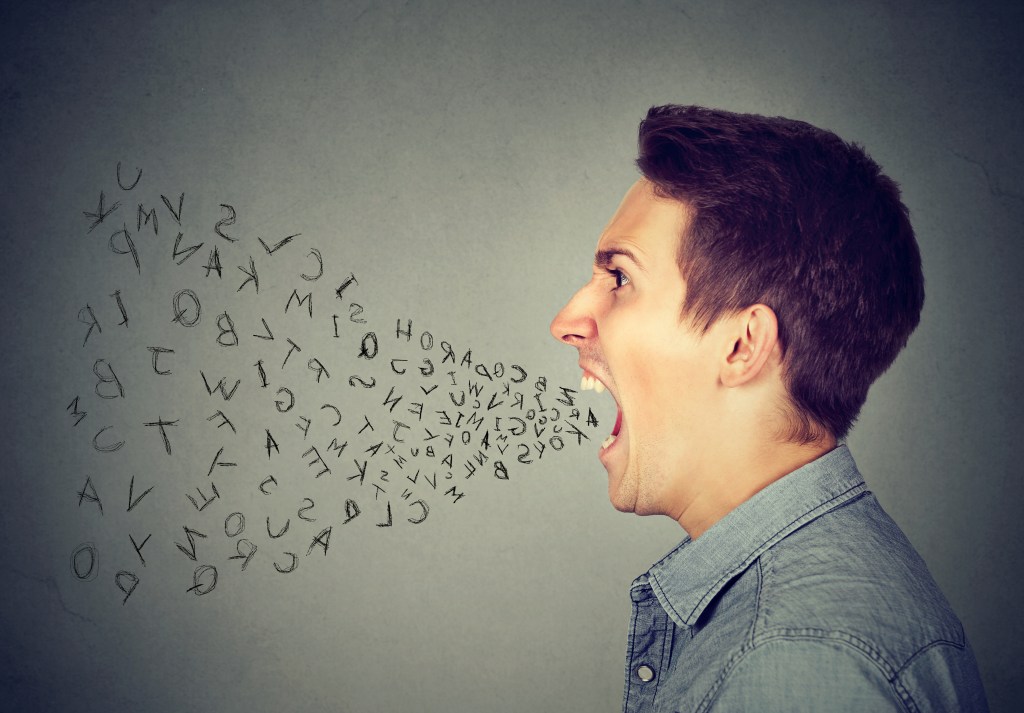 Side profile portrait of young angry man screaming with alphabet letters flying out of wide open mouth isolated on gray wall background.